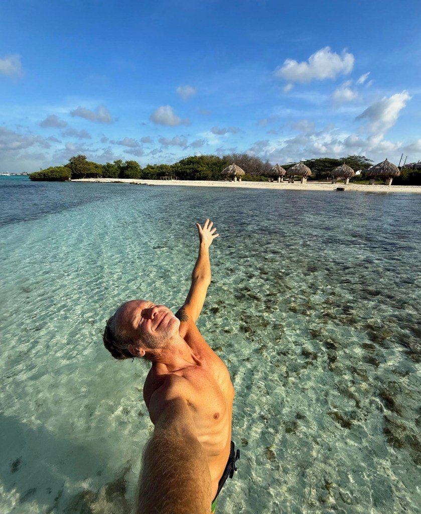 Stephen Lashbrook standing in clear blue water with his arms raised, smiling.